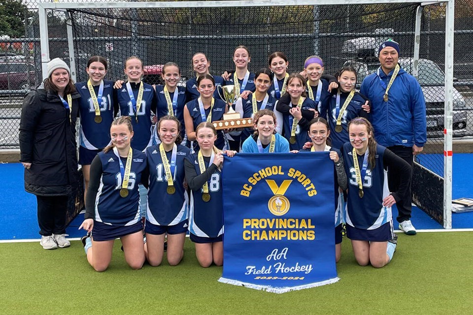 A girls field hockey team pose together adorned with medals, a trophy and banner.