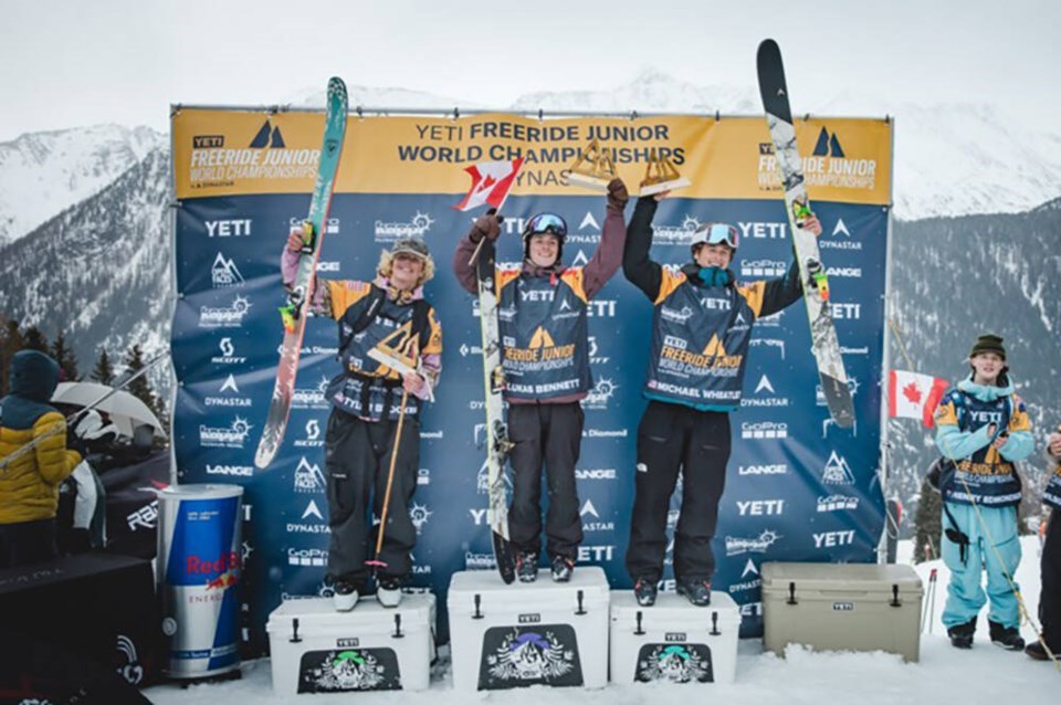 Three male skiers stand on an outdoor podium holding their skis and trophies in celebration.