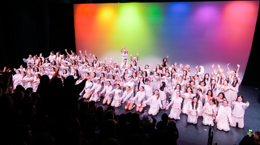 A wide view of a full ensemble of performers dressed in white costumes gathered on stage beneath a rainbow‑coloured backdrop, wi
