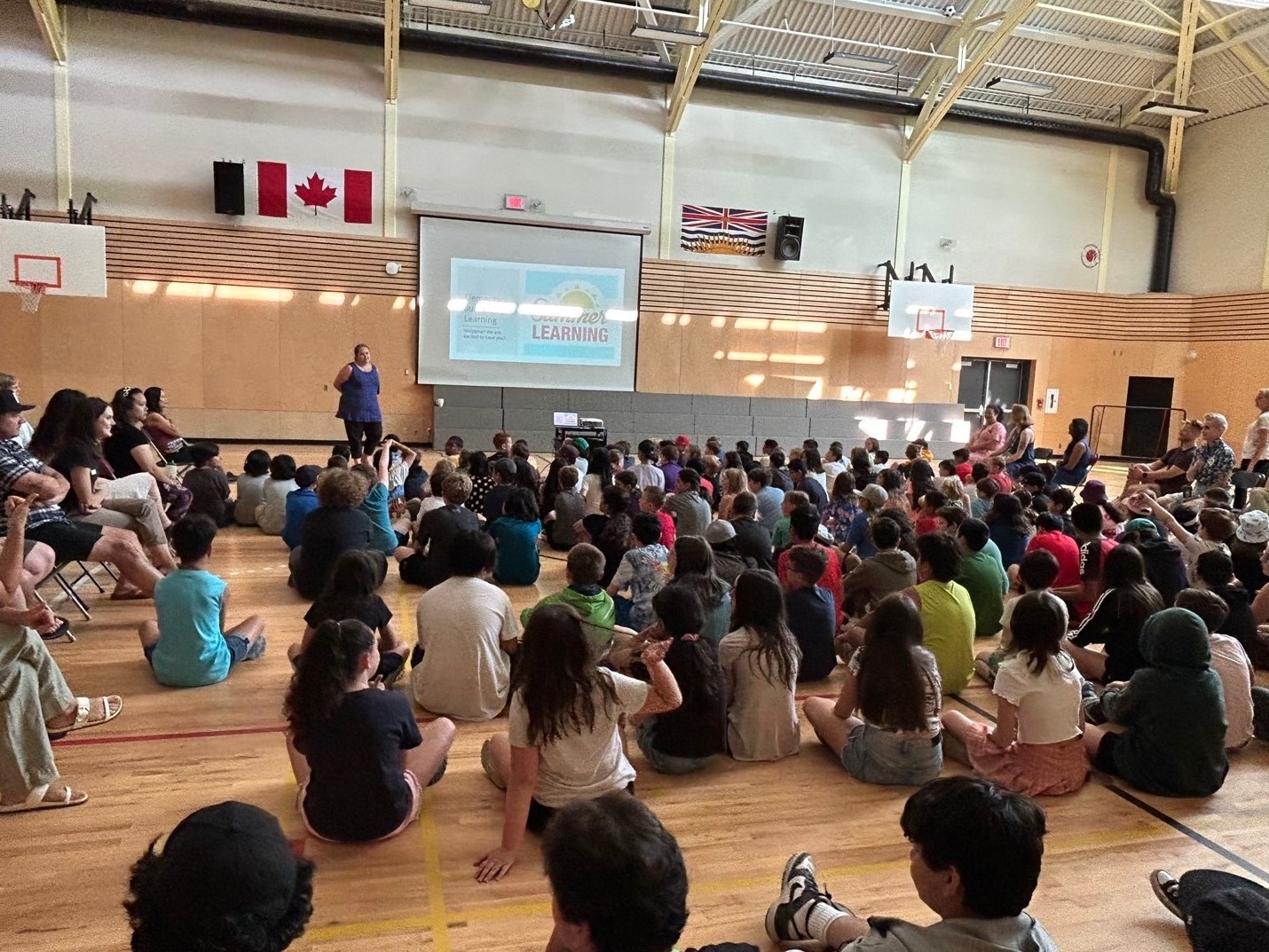 Students gather sitting on a gym floor looking at a projected screen that says Summer Learning.