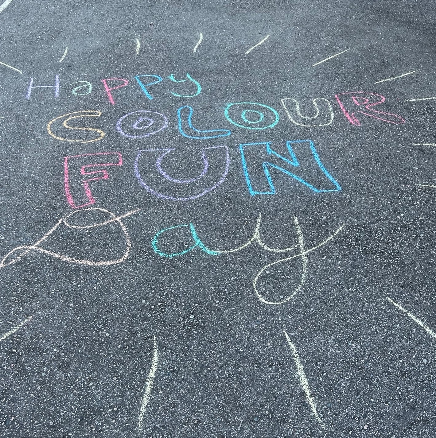 Happy Colour Fun Day is written in mulitcolour chalk on pavement.