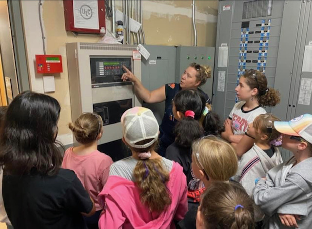A group of young girls watch a female educator talk about an electrical panel.