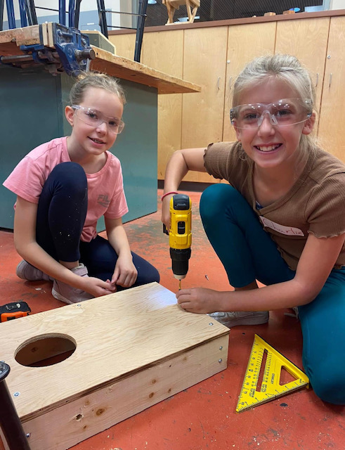 Two students wear safety glasses and use a drill and corner to build a rectangular box out of wood.