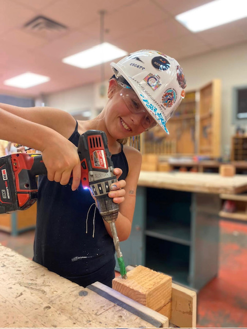 A young student wears a white hard hat covered in stickers while using a drill in a block of wood.