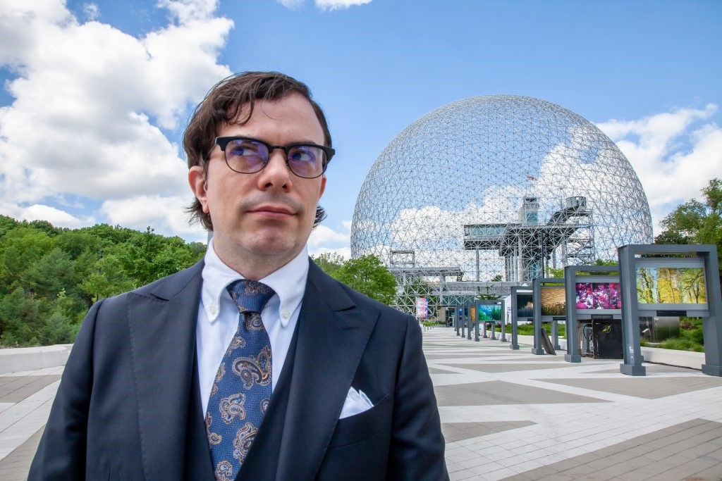 Darcy James Argue wears a blue suit and glasses. Located outside in a courtyard with a geodesic dome.
