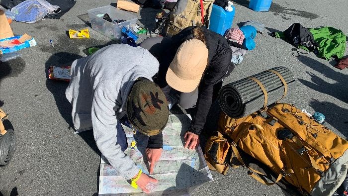 Two people hunch over a map placed on concrete. They are surrounded by camping gear.