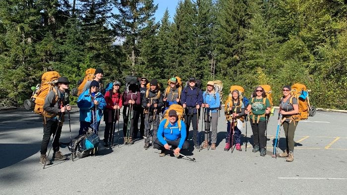 14 people stand with packed hiking bags and poles while smiling towards the camera.