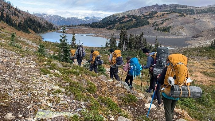 A group of people hike in single file in a sub-alpine meadow. A small lake is in the distance.
