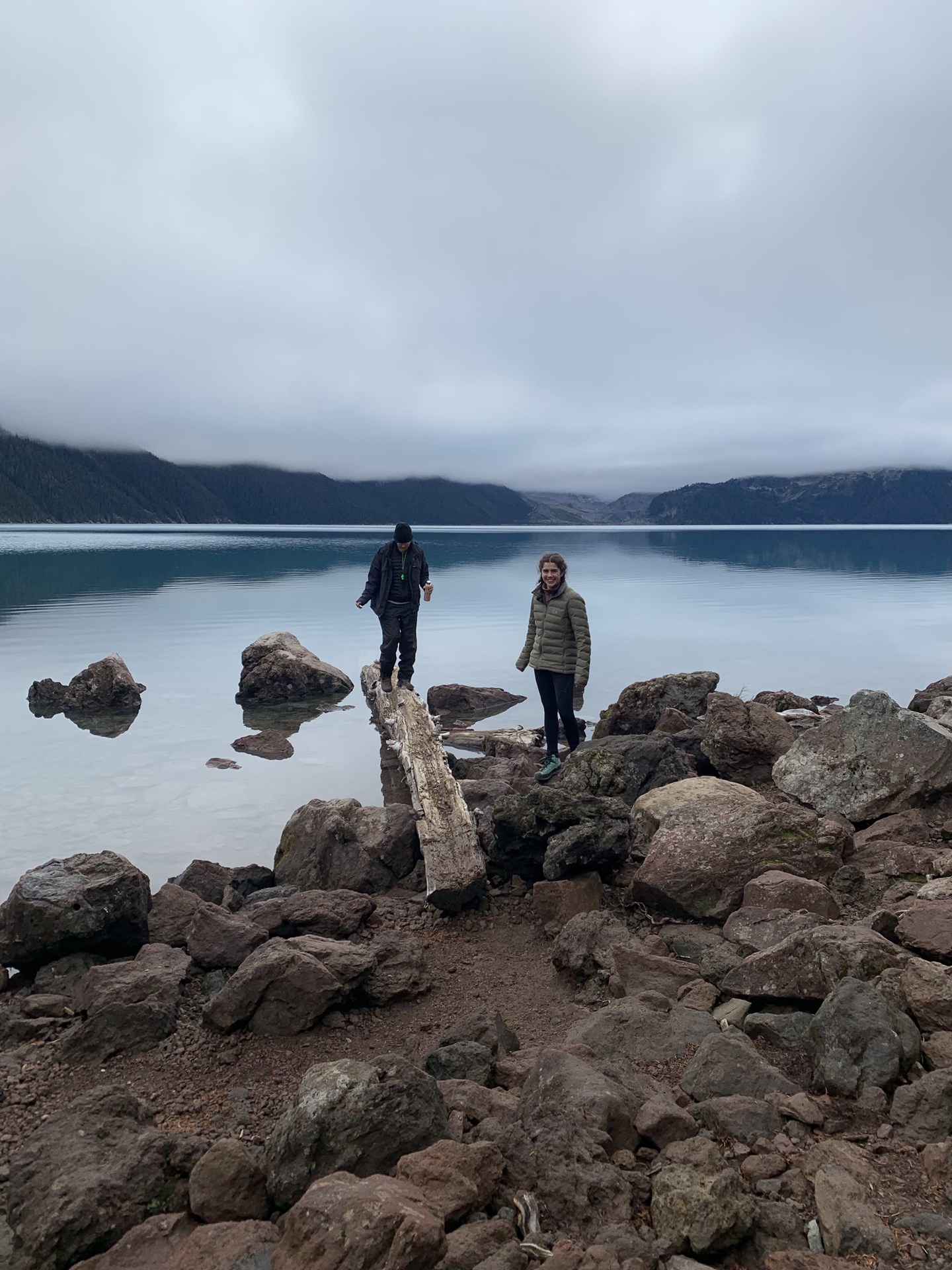 Two people stand on the shore of a still lake. The sky is overcast.