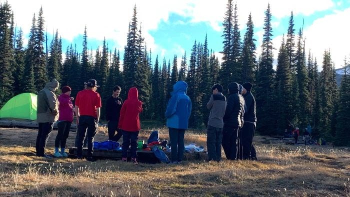 Surrounded by trees, a group of people stand around camping gear at a campsite. Early morning sun shines from the right.