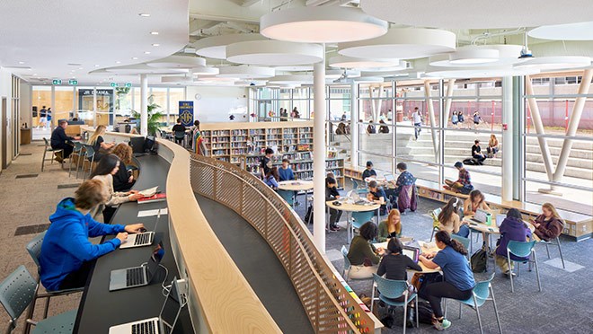 Wide angle photograph of the inerior of a library with students working at various tables.