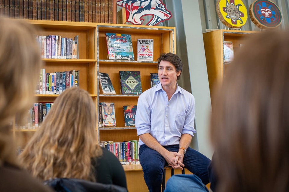 Prime Minister Justin Trudeau sits on a stool in front of a library book case speaking to a room full of students.