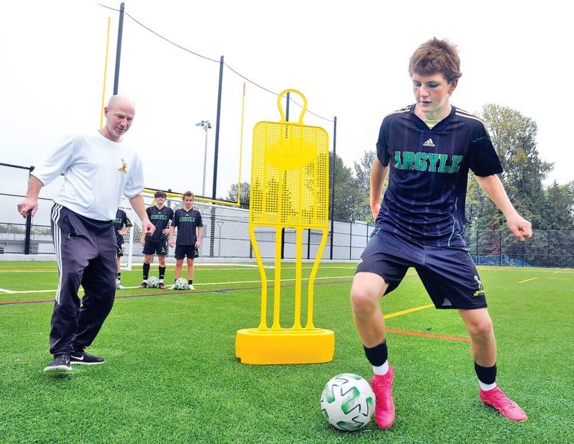 A soccer team member and coach take part in a drill on an artificial turf field.