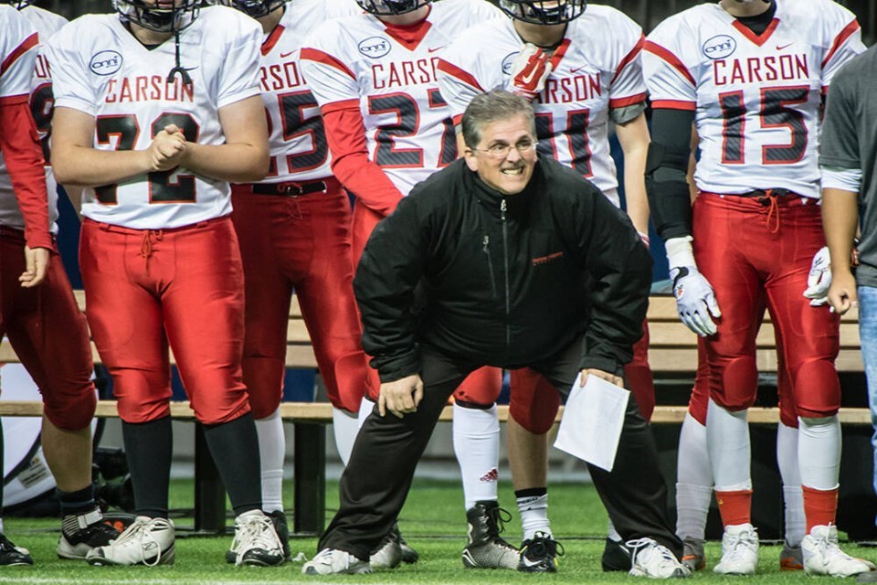 A coach dressed in an al black tracksuit braces himself with his hands on his knees. Football players stand in line behind him.