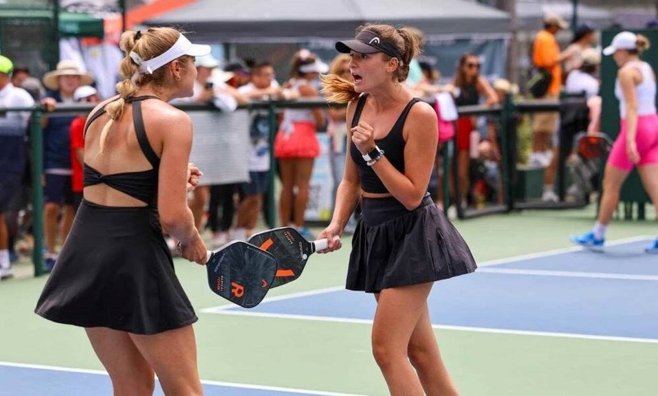 In a moment of celebration two girls tap paddles while holding their other hand in a fist. They are both wearing black outfits.