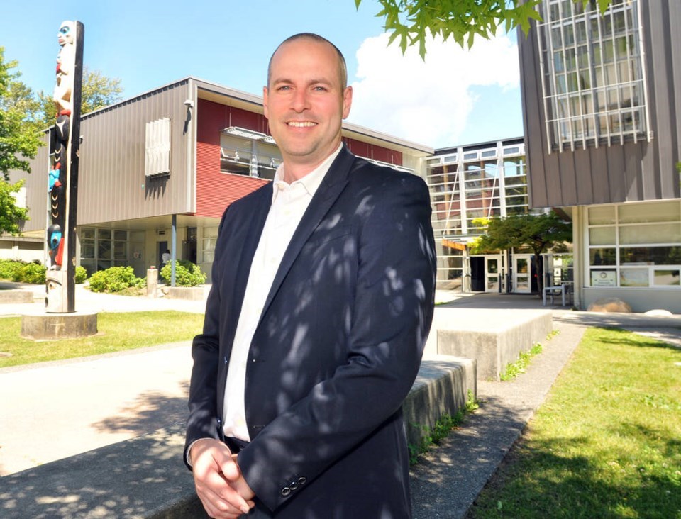 Person in a dark suit standing outside a modern building on a sunny day, with a totem pole and trees nearby.
