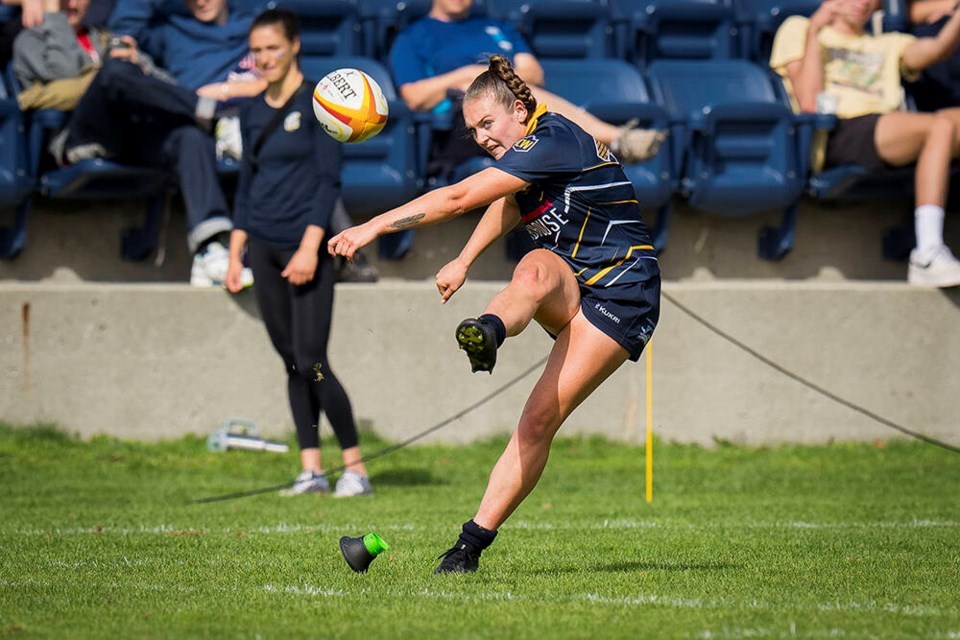 Savannah Bauder kicks a rugby ball on a brigt green field on a sunny day. Seats behind her are filled with an audience.