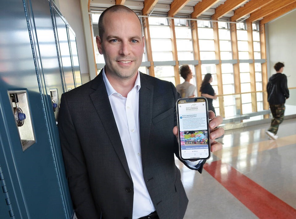 Principal Mark Barrett smiles to the camera while leaning against tall blue lockers while holding a phone screen to the camera.