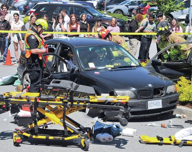 A crashed black car with medical and fire personnel attending the passengers. Students view from behind caution tape.